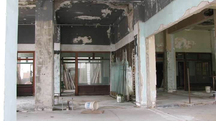 An interior room in a decayed condition, with bare concrete block and missing paint all over. Wooden paneled windows are visible in the back.