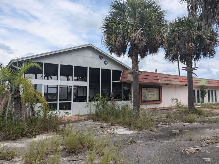 A one story building with a patio-style addition on the side. 3 palm trees stand in front and tall grass grows through the cracked asphalt in multiple places