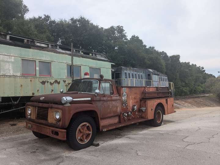 a small firetruck from the 1960s sits in front of the passenger car and locomotive. It's entirely covered in shades of dark umber to orange rust