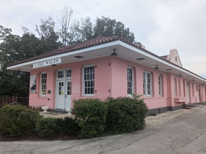 A 3/4 view of a railroad station building, one story, painted pink with white trim, doors, and windows, and dark red 'barrel' clay roof tiles