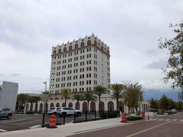 A 10-story stucco hotel building, standing over a parking lot in the foreground. There's several palm trees along the street in front