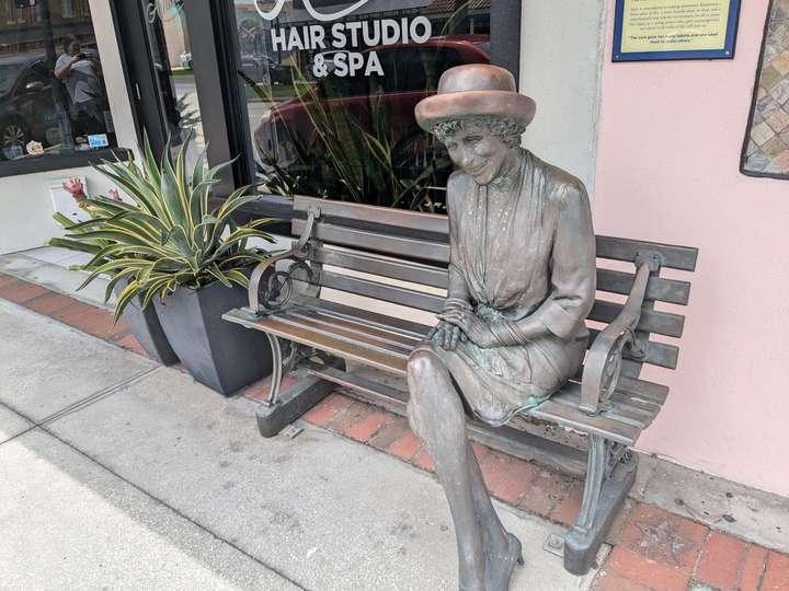 A bronze statue of a smiling elderly woman sitting on a bench with her legs crossed