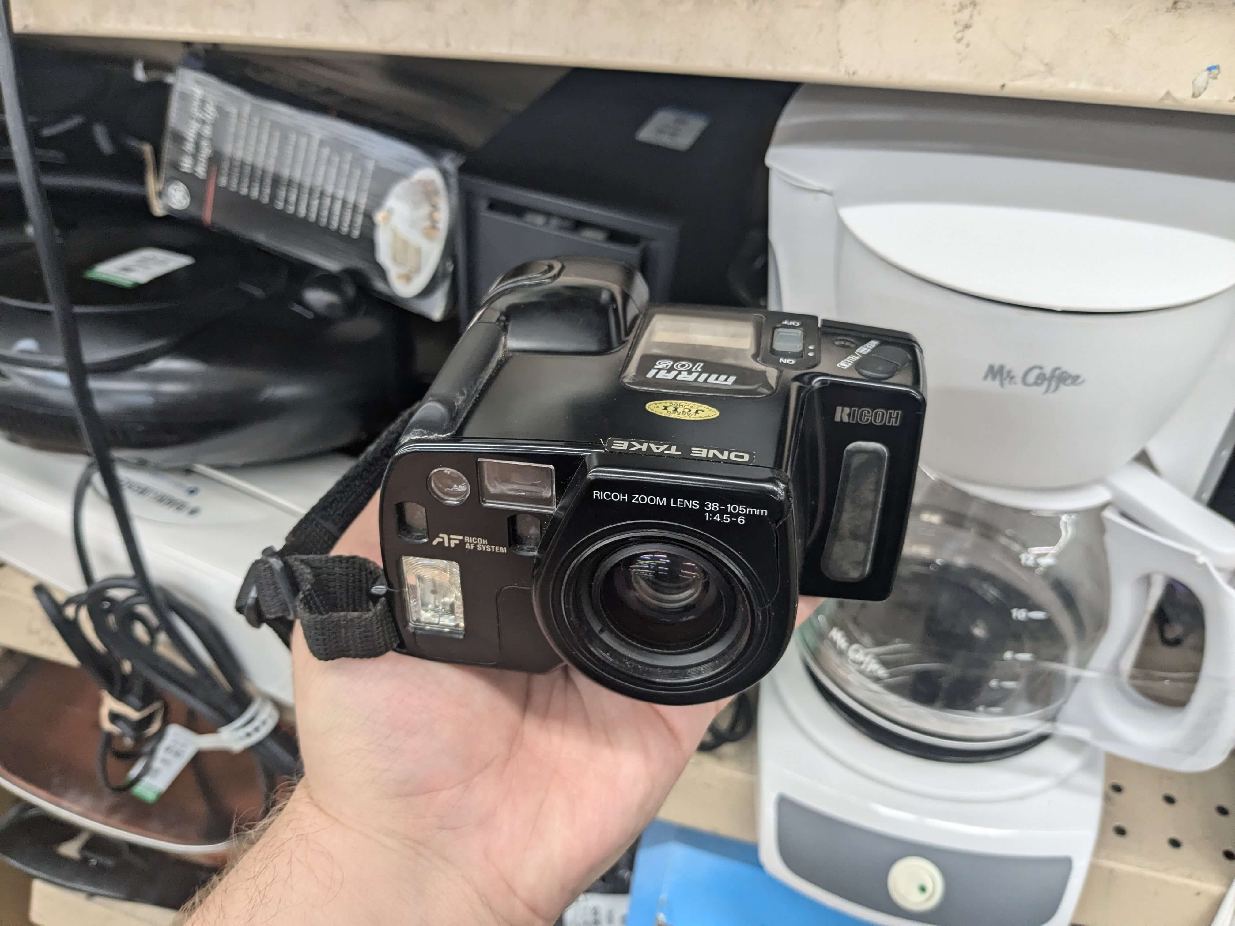 an oddly shaped 35mm film camera, held in front of a shelf of thrift store electronics