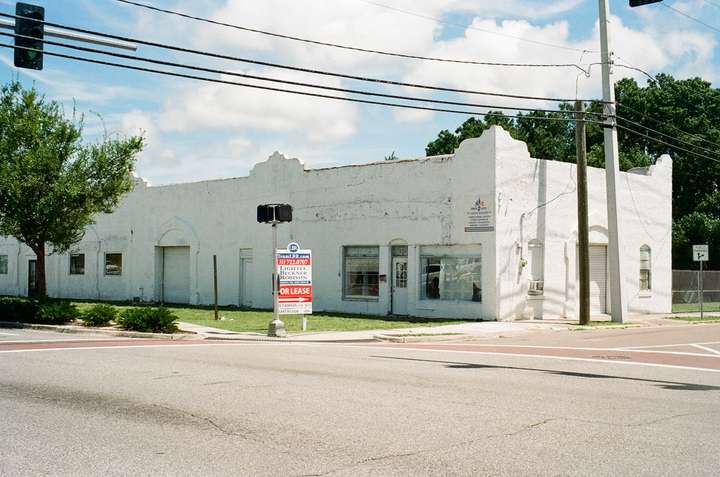 A long stuccoed building on a street corner, with a For Lease sign in front