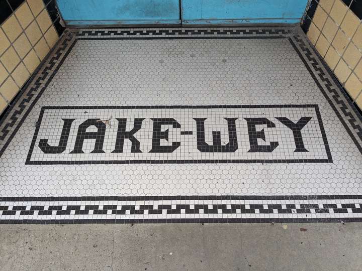 a tile entryway with serifed letters spelling out JAKE-WEY, surrounded by a thin outland. a lacey pattern surrounds the whole entrance