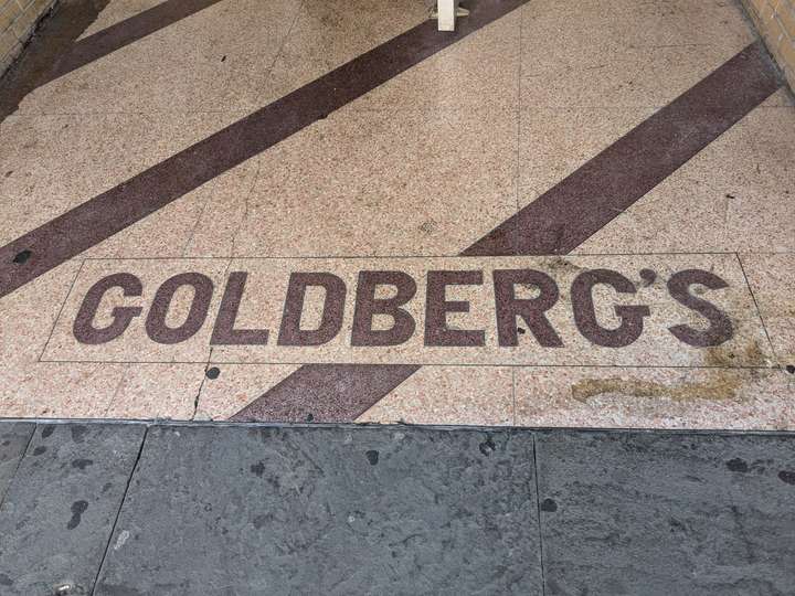 GOLDBERG'S in dark red, block font, cut with diagonal stripes of the same color against a sand yellow background