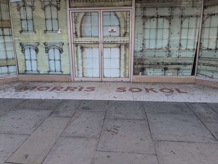 A wide shot of an entryway that reads MORRIS-SOKOL in dim red letters, against a speckled white background. Artwork depicting a building covers the windows behind