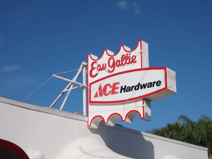 A red and white sign with scalloped top and bottom edges, and a protruding middle. Eau Gallie is spelled in neon script text, with Ace Hardware on a simple plastic panel underneath