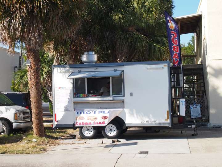 A mostly white enclosed food trailer with a banner hanging underneath the serving window advertising Cuban food