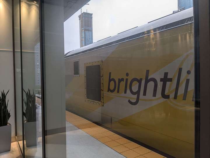 A train viewed at an angle through a window; it's in a yellow and white livery that says Brightline