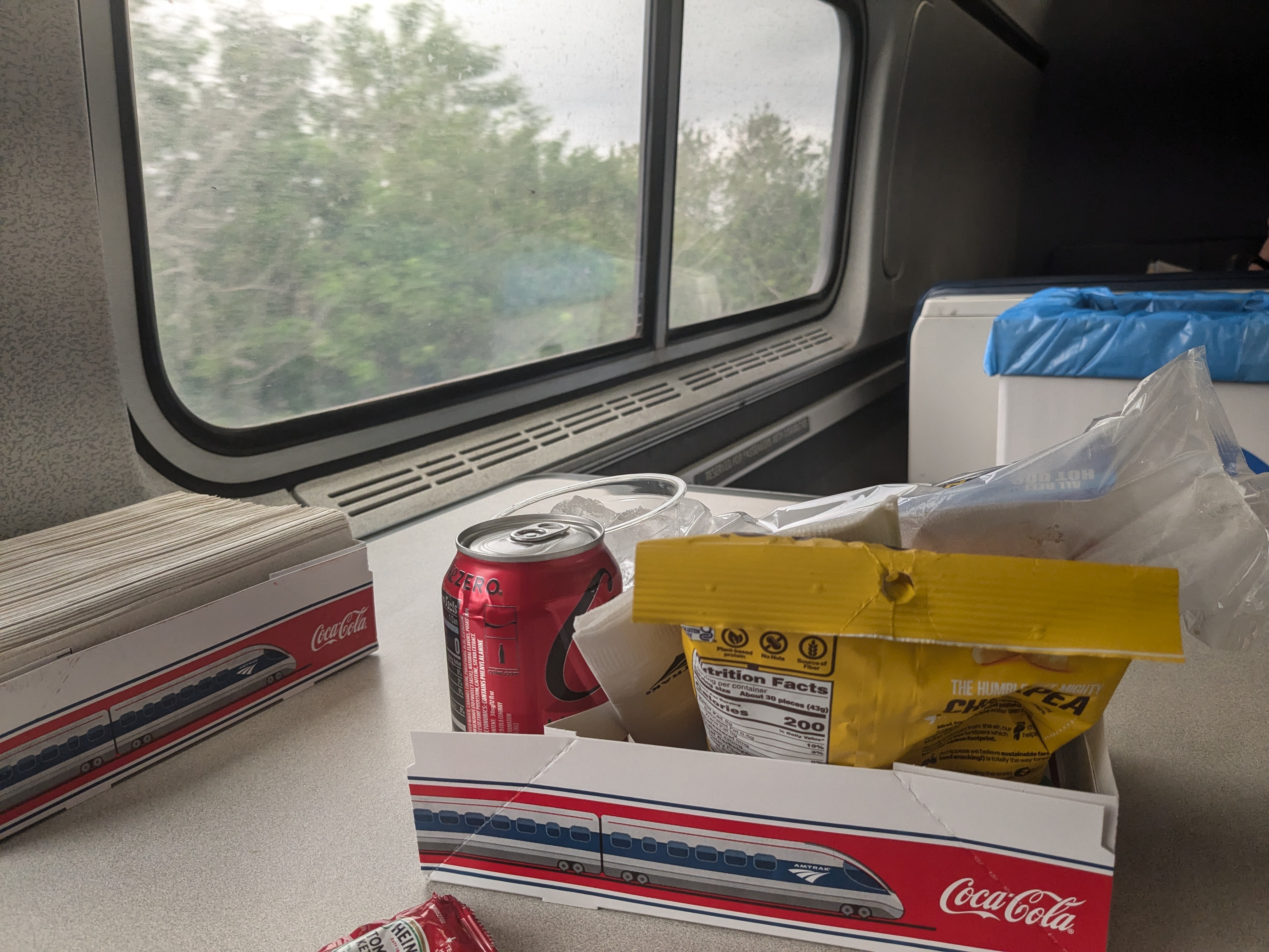 A boxed lunch of hot dogs, chips, and a canned soda sit in the foreground on a table. In the background, trees are seen passing by through a window