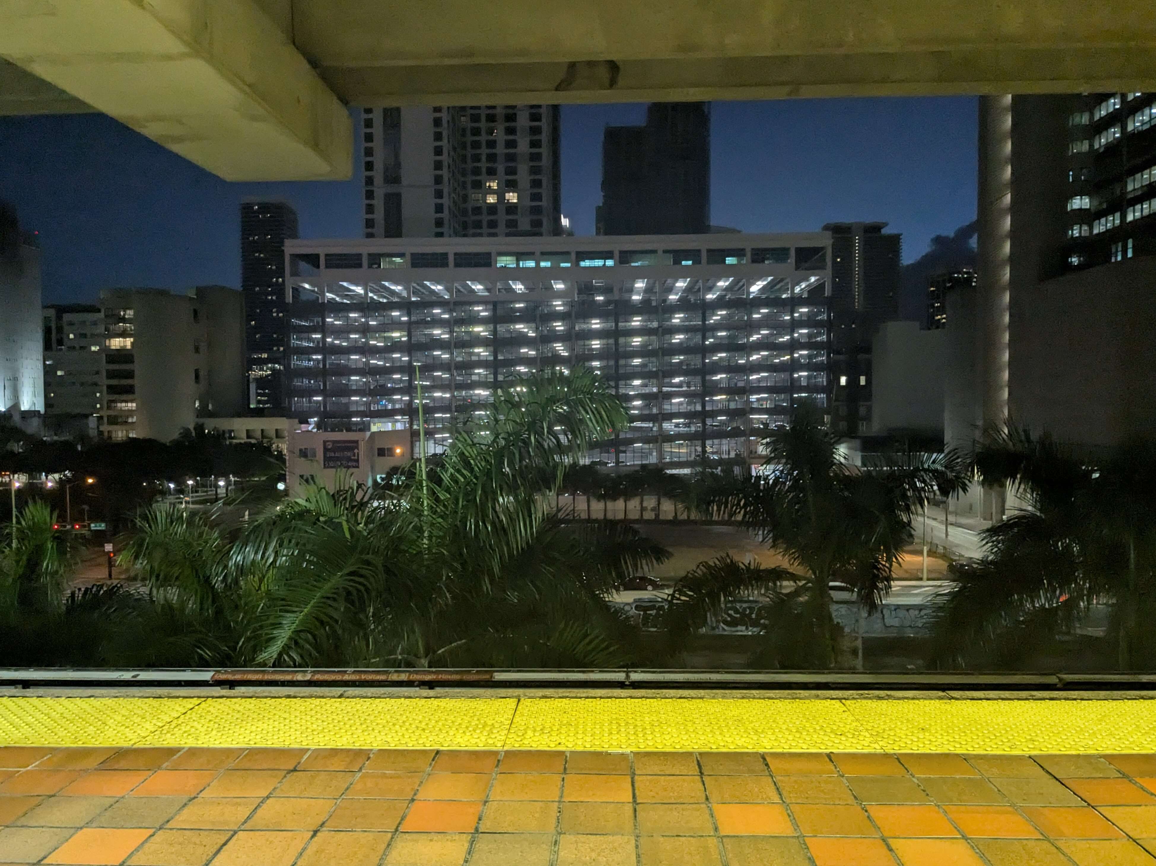 A view of palm trees and a large, brightly lit high rise parking garage, against an urban skyline and the night sky