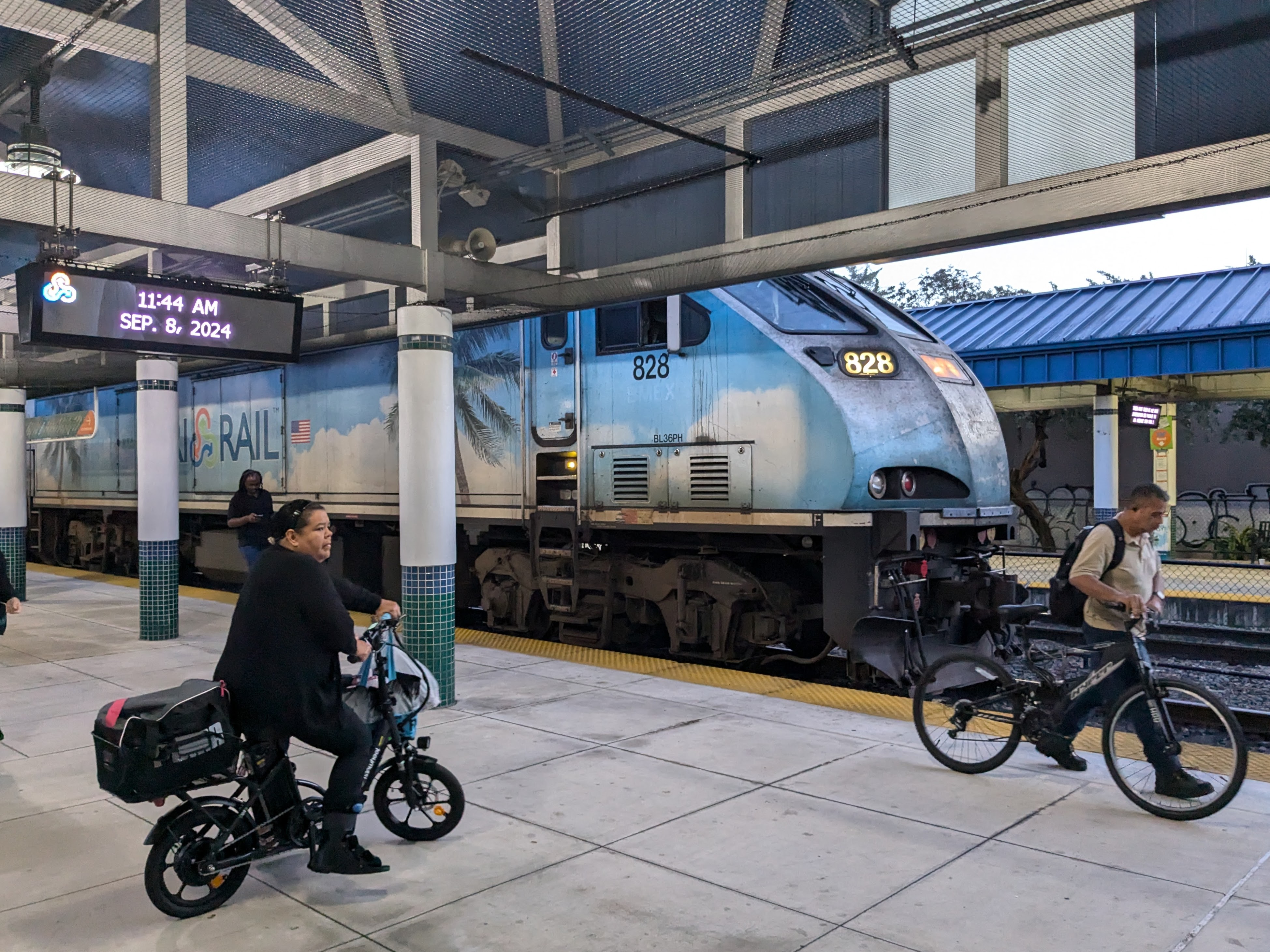 A blue streamlined diesel locomotive sits next to a station platform. On either side in the foreground, commuters are leaving with their bikes