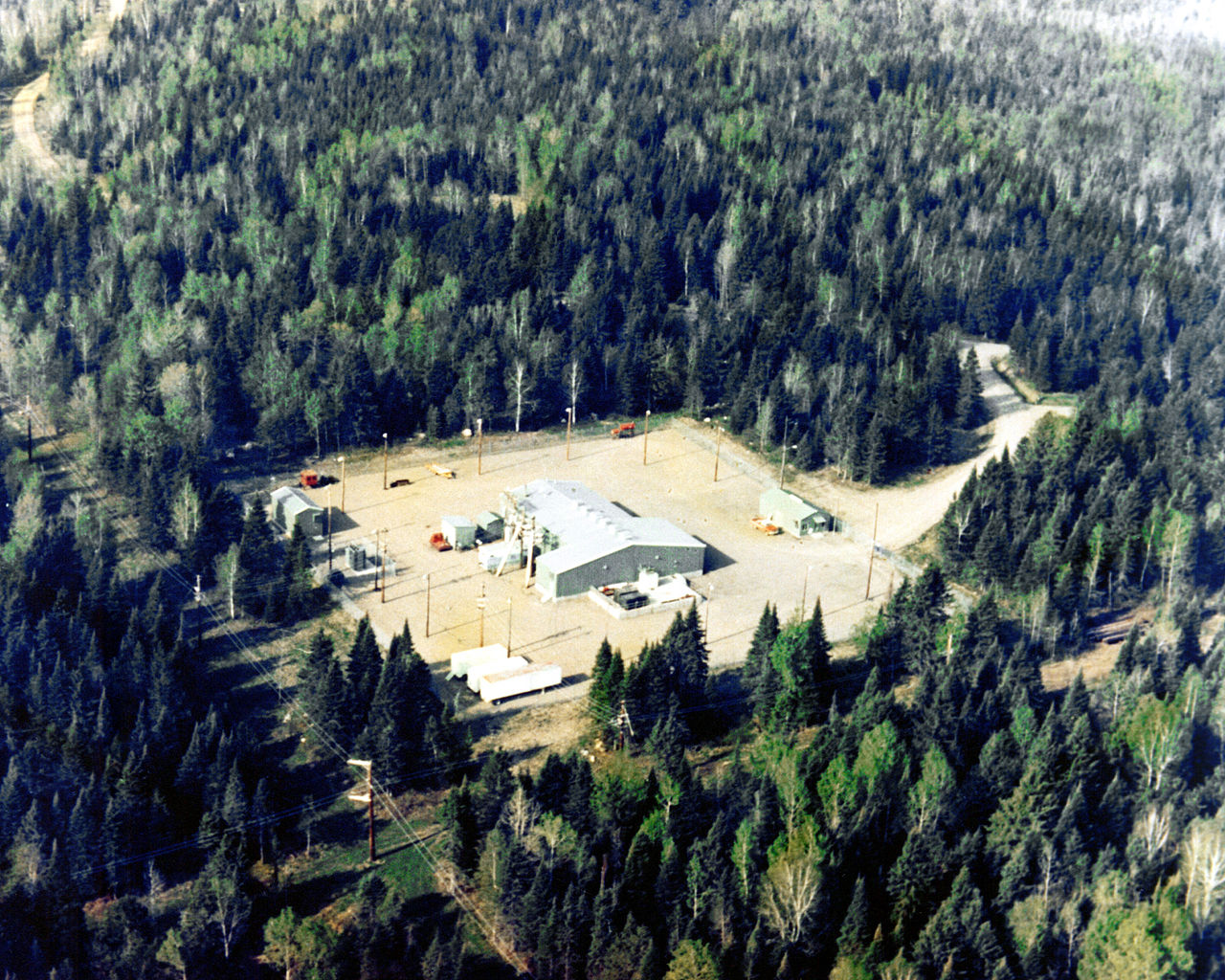 An aerial photo of a clearing in a thick forest, with one medium sized metal building and a few smaller sheds surrounding. Cables strung between poles are visible in the surrounding area.